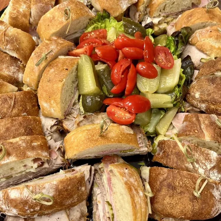 A catering platter of halved sandwiches around a bowl of pickles and cherry tomatoes